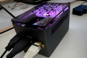 Ein Raspberry Pi, eingebaut in ein kleines, schwarzes Gehäuse mit Lüftungsschlitzen und einem transparenten Deckel, durch den violette Beleuchtung sowie das Innenleben ein wenig zu sehen sind. An der Seite sind mehrere Kabel angeschlossen.