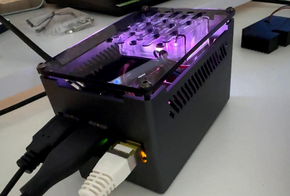 Ein Raspberry Pi, eingebaut in ein kleines, schwarzes Gehäuse mit Lüftungsschlitzen und einem transparenten Deckel, durch den violette Beleuchtung sowie das Innenleben ein wenig zu sehen sind. An der Seite sind mehrere Kabel angeschlossen.