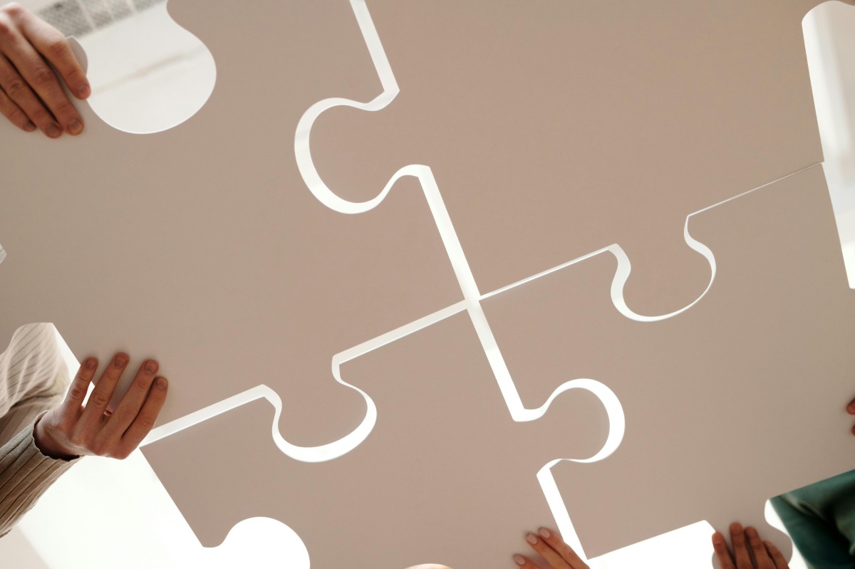 Das Bild zeigt drei Personen, die gemeinsam ein großes Puzzle auf einem Tisch zusammensetzen. Die Hände der Personen sind sichtbar, während sie die Puzzleteile halten und platzieren. Das Puzzle besteht aus mehreren großen, beigefarbenen Teilen mit weißen Konturen.